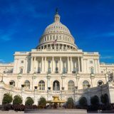Front facade of the U.S. Capitol with its dome, flag, and steps in the foreground against a blue sky.