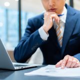 Businessperson in a blue suit reviews documents beside an open laptop on a desk. Transport of focus is on paperwork and workspace.