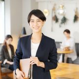 Smiling businesswoman in a navy blazer, holding a tan notebook in a bright cafe setting with coworkers blurred in the background