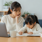 Mother watches her young daughter as she writes in a notebook at a table, with a laptop and a mug nearby.