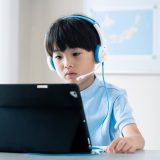 Young boy wearing blue and white over-ear headphones uses a tablet on a stand for learning in a classroom.