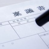 Close-up of a hand with a pen writing on a Japanese tax return form (確定申告) on a desk surface.