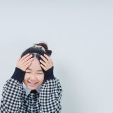 Childhood portrait of a smiling girl covering her head with both hands, wearing a black-and-white checkered coat, against a light gray wall