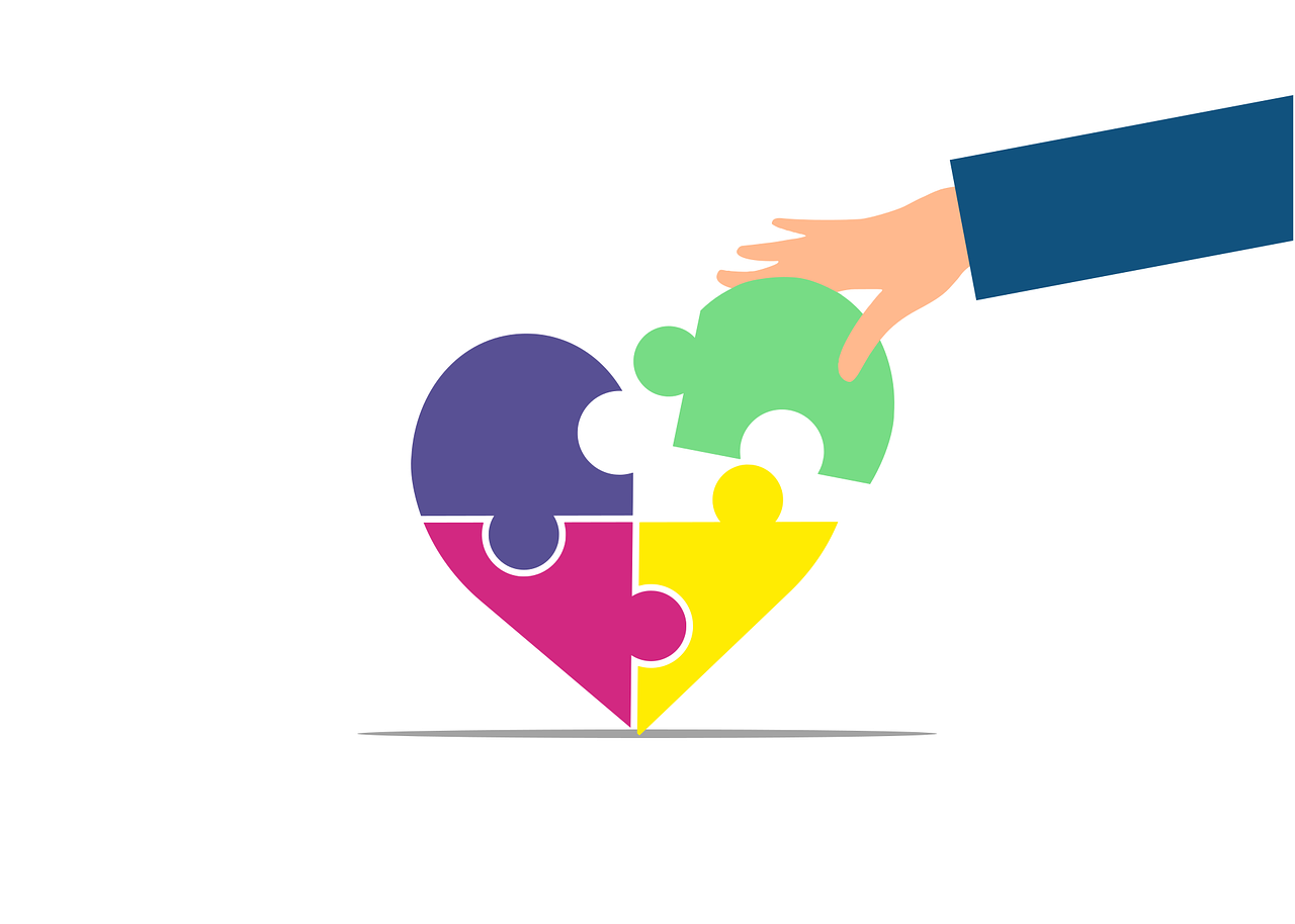 Hand placing a green puzzle piece into a multicolor heart made of puzzle pieces, symbolizing teamwork and support.