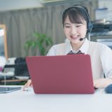 Woman with headset smiling while working on a pink laptop at a desk in an office.