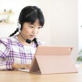 Girl with a headset using a tablet at a wooden table, typing on a keyboard in a bright room.