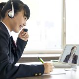 Young woman in a dark blazer wearing a headset, smiling during a video conference at a bright desk with a tablet showing a colleague on screen.