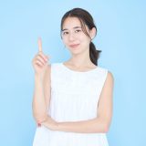 Portrait of a woman in a white sleeveless dress against a blue background, raising her index finger.