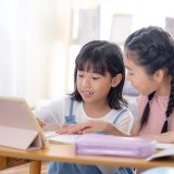 Two young girls sit at a table, looking at a tablet while writing in a notebook.