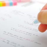 Close-up of a hand writing with a blue pen on a sheet, with colorful highlighters blurred in the background