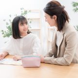 Girl writing at a desk while a woman mentor leans in, smiling; a pink pencil case sits nearby in a bright room with plants in the background.